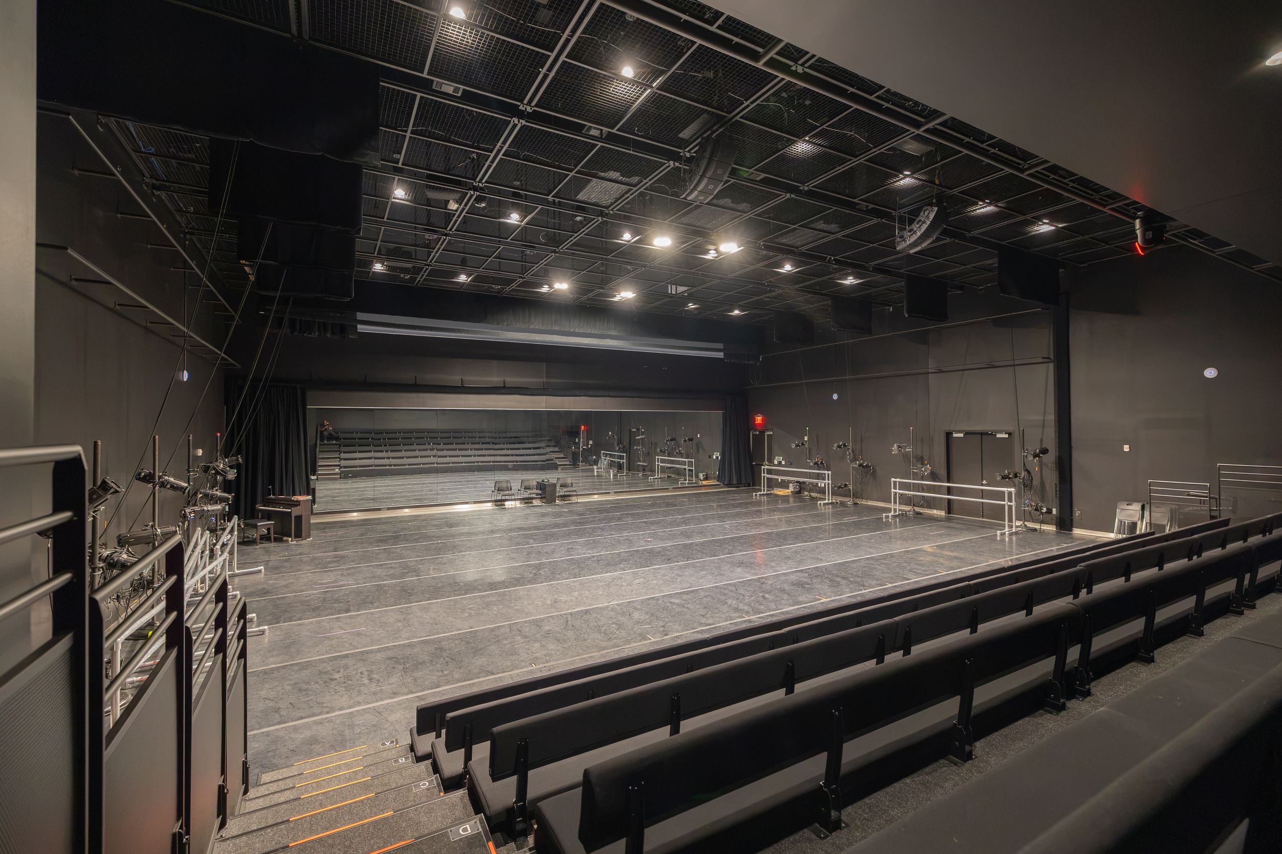 View of large black-box rehearsal space with seating on risers and bright overhead lights.