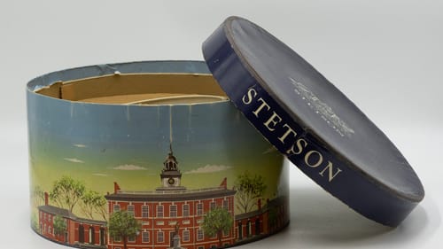 Vintage round hatbox decorated with a painting of an old brick building, Stetson written on its top, leaning against the box