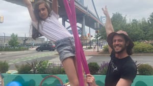 A man helps a young girl on an aerial trapeze fabric outdoors, the Ben Franklin bridge in the background