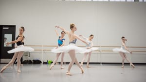 Five ballet dancers in white tutus, cream colored tights and varying designed/colored leotards practicing in a rehearsal room