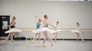 Five ballet dancers in white tutus, cream colored tights and varying designed/colored leotards practicing in a rehearsal room