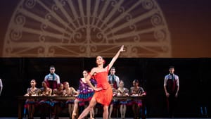 A dancer in an all red dress poses on stage in front of other dancers, an image projected on a screen behind them