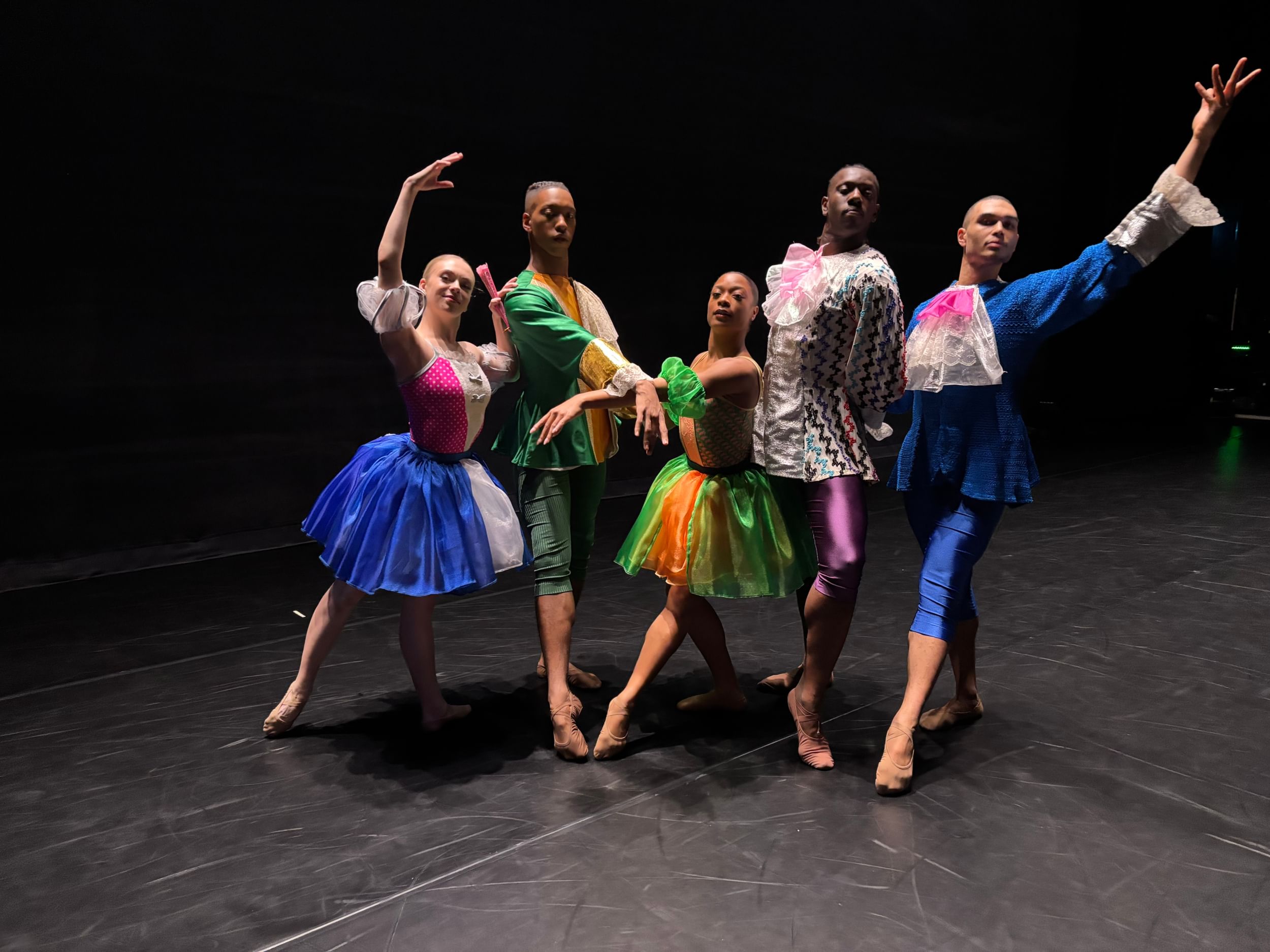 Five dancers with different skin tones pose together, limbs extended, in colorful frilled costumes reminiscent of the 1600s