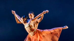 Two Black dancers in pas de deux on a dark purple tinted stage in red see-through outfits