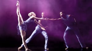 A woman dancer with one leg extended up pulls against two male dancers who lean toward her, grabbing her hands.