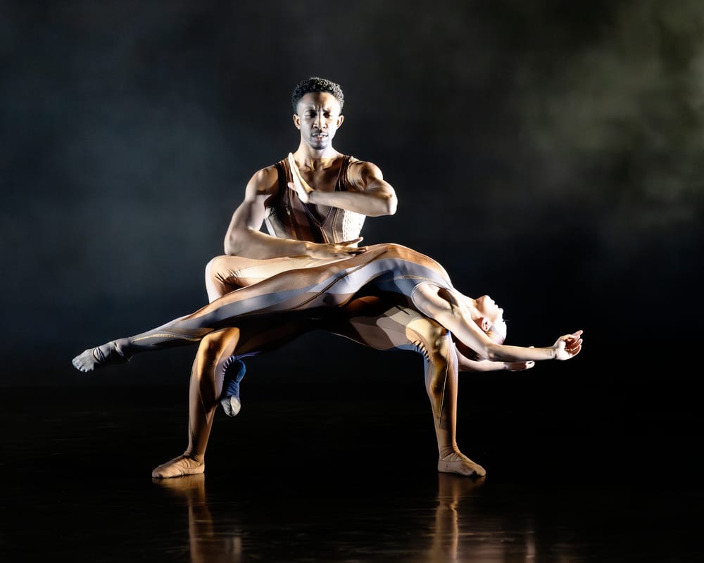 Gilbert squats with Fenton lying gracefully extended across his knees. They both wear bronze, silver & gold bodysuits.