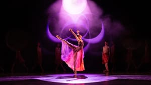 Under dramatic orbs of misty white & purple light, a dancer in a filmy dress poses en pointe with leg and arms outstretched