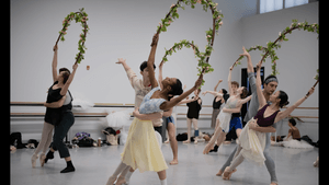 Five pairs of dancers in rehearsal, all in pas de deux, the women holding large rose vine garlands.