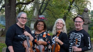 Four players with their violas dressed in Halloween-themed tops pose for a group photo in a graveyard during daytime.