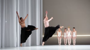 Two male dancers in long black skirts jump high in unison, and four women dancers stand in white leotards behind them.