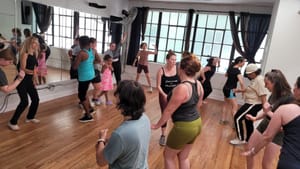A multi-racial group of about 17 dancers of all ages dance together in a small, windowed studio with wooden floors.