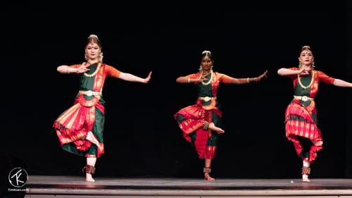 Three female dancers in green and red outfits strike the same pose on one foot, left arm extended, in a South Asian style