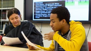 Two teenagers, one in a black hoodie and the other in a yellow hoodie, sit at a table side by side reading a script aloud.