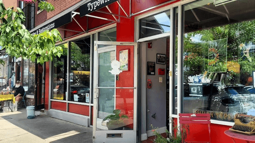 Summer day photo of the storefront with door open. It has large glass windows, a red front, and a black awning.