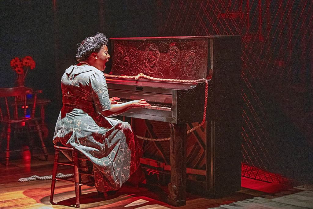 With her back to the viewer, Johnson, in a 1930s era dress, sings and plays an elaborately carved wooden piano.