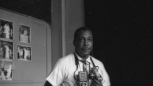 Withers, a Black man, holds two film cameras, and poses for a b&w portrait. His photos are behind him on a wall.