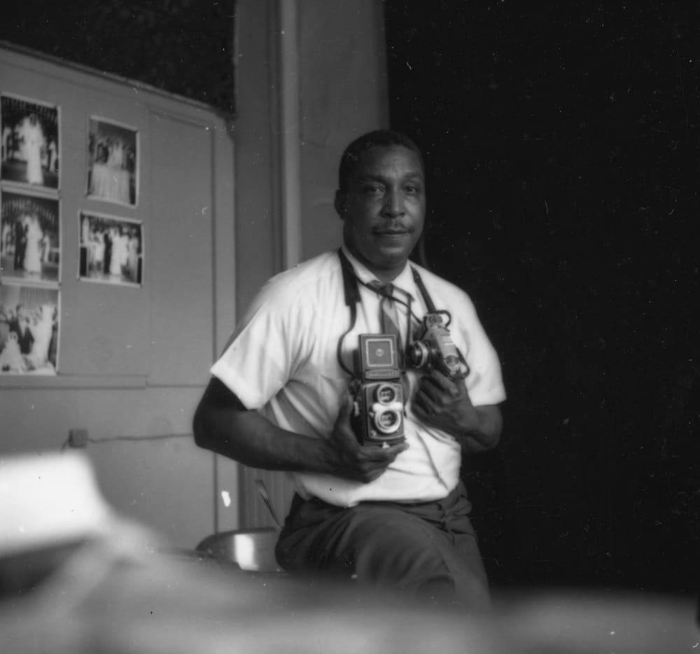Withers, a Black man, holds two film cameras, and poses for a b&w portrait. His photos are behind him on a wall.