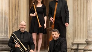 The four players in all black pose outdoors on stone steps in front of a door, holding shawms and trombones