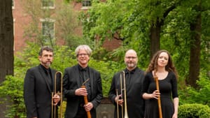 The four in the ensemble pose outside in a public-park-like daylit area, wearing all black, holding wind instruments