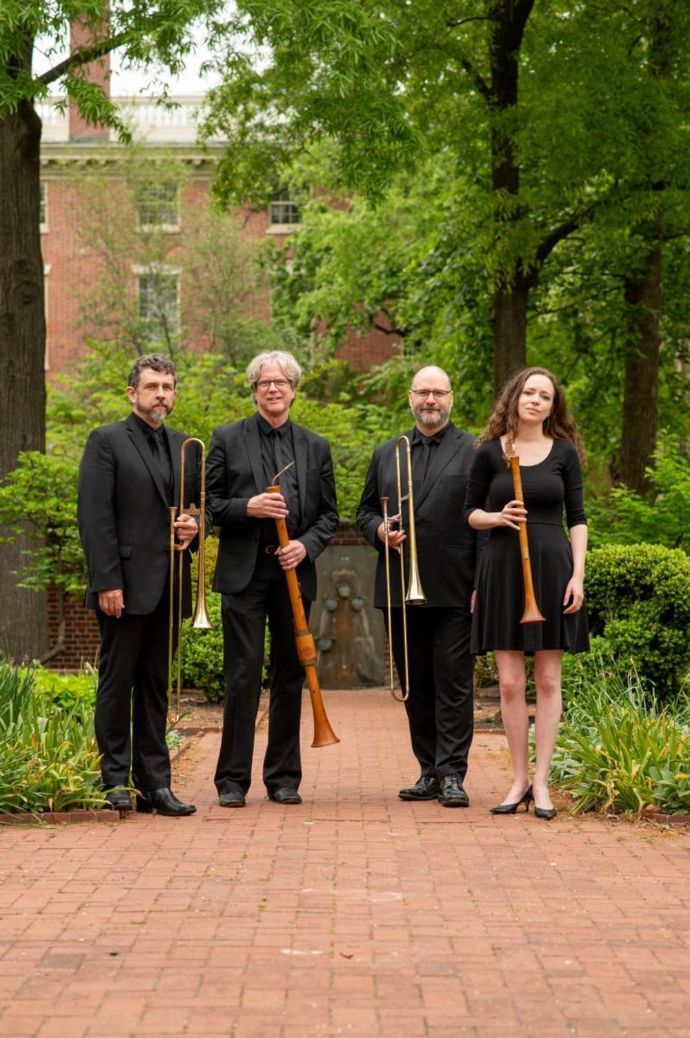 The four in the ensemble pose outside in a public-park-like daylit area, wearing all black, holding wind instruments