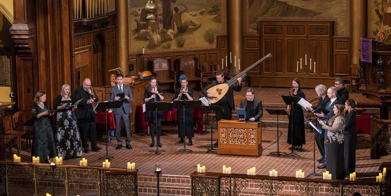 14 period musicians and singers in formalwear perform in a semicircle on a church chancel with a brick floor & wood paneling