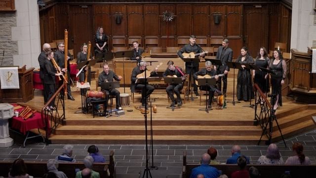 15 singers & musicians on Renaissance-era instruments perform together on a wooden chancel.