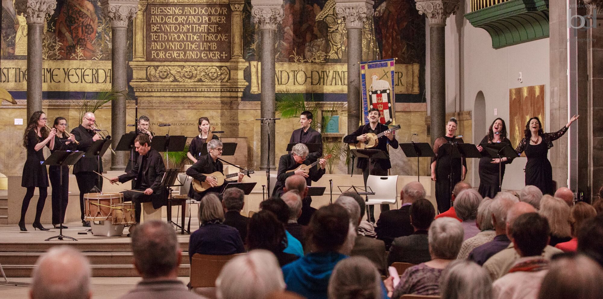 On a pillared chancel with a golden mural backdrop, 13 musicians and singers, all dressed in black, perform.