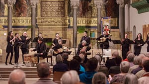 On a pillared chancel with a golden mural backdrop, 13 musicians and singers, all dressed in black, perform.