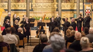 13 singers and musicians, all dressed in black, perform on the gold-painted, pillared chancel of a church full of listeners.