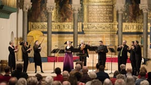 Rottsolk, in a black & pink dress, sings on a golden, pillared chancel with 8 black-clad musicians playing around her.