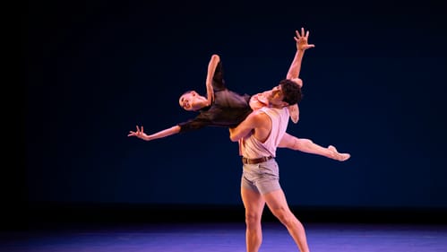 Two dancers on a stage dimly lit with purples and pinks. One dancer holds up another, who extends out long, both expressive