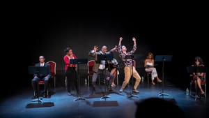 A diverse cast of seven, some seated and some standing and gesticulating wildly, perform on a black stage with music stands