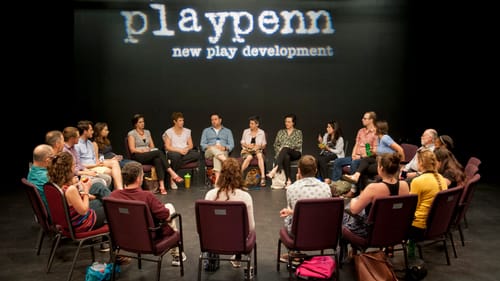 22 people in casual clothes sit in a circle of chairs on a stage under the projected PlayPenn logo.