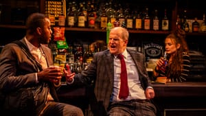 Akintilo and Lawton talk animatedly at a bar with rows of liquor bottles. Congdon sips a beer, looking annoyed behind them
