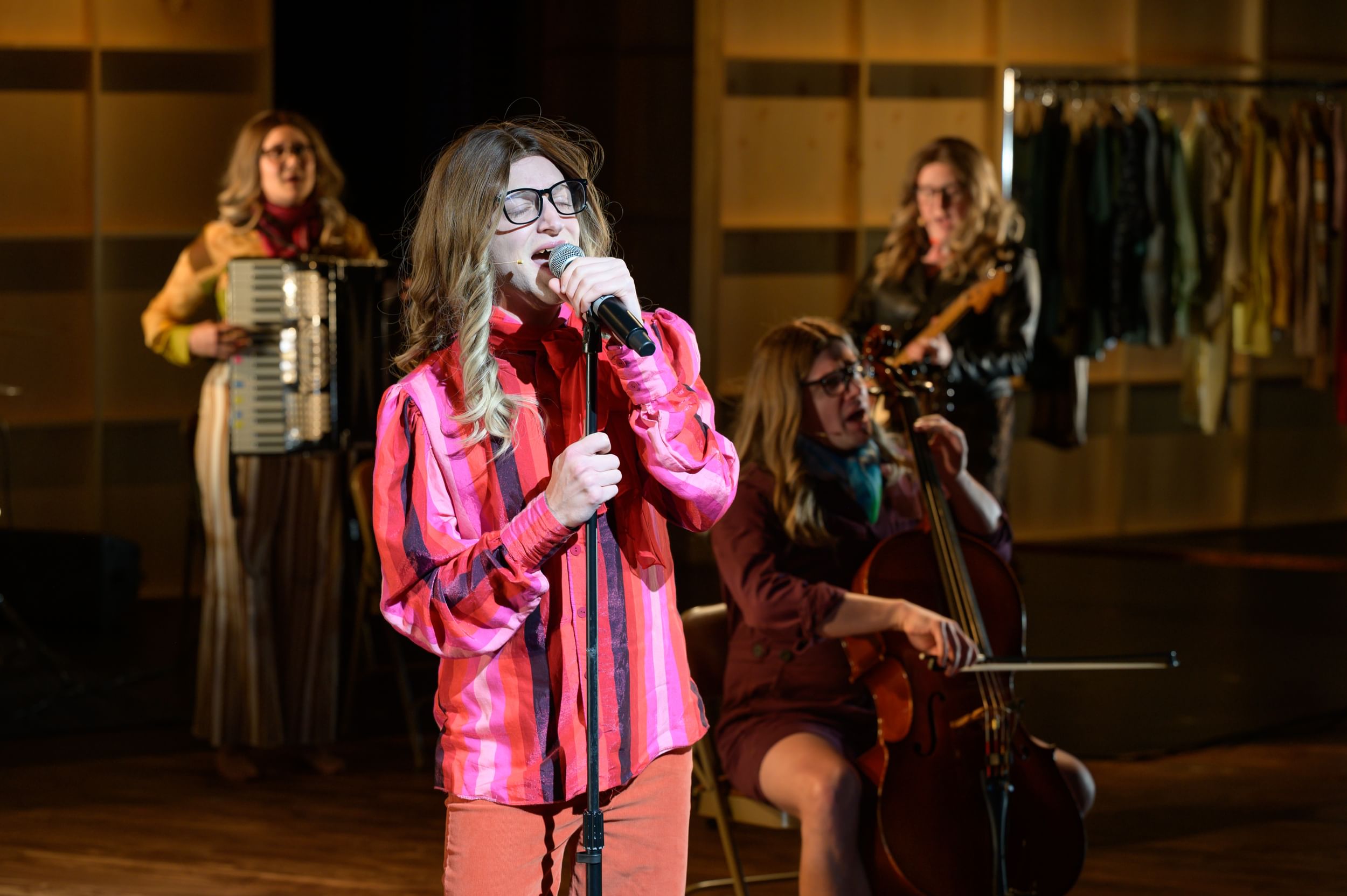 Ressler, a white person in a blonde wig & pink-striped shirt, sings into a mic with three musicians in the background.
