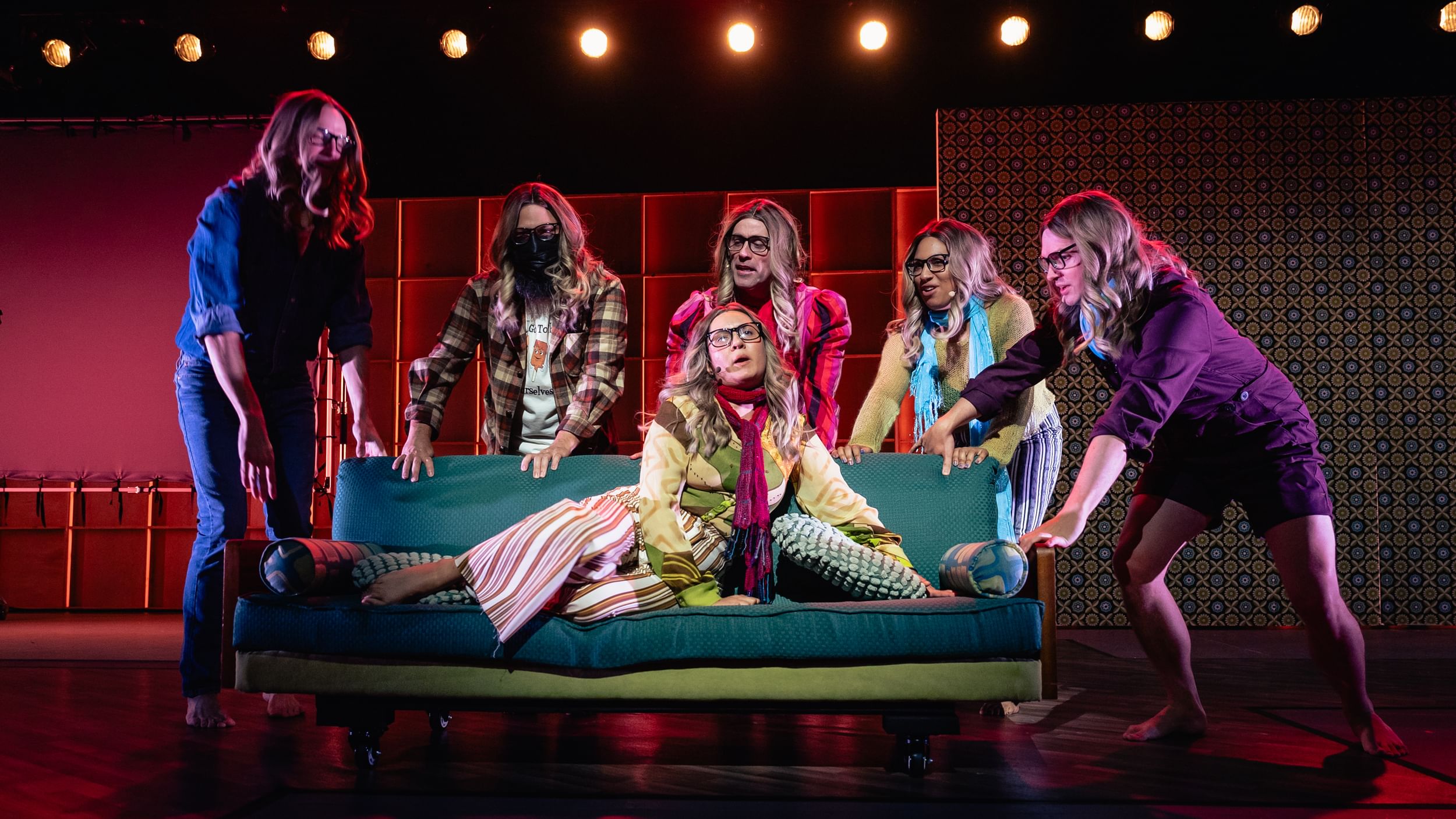 Six performers on stage all wearing similar blonde wigs and glasses sing, one on a small couch, others stand behind