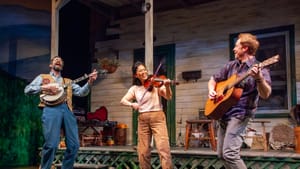 Three performers play on a porch on a stage, one with a banjo, a violin, and an acoustic guitar