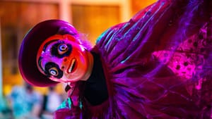 A person dressed in a see-through, flowing robe wears a smiling black, red, and white mask