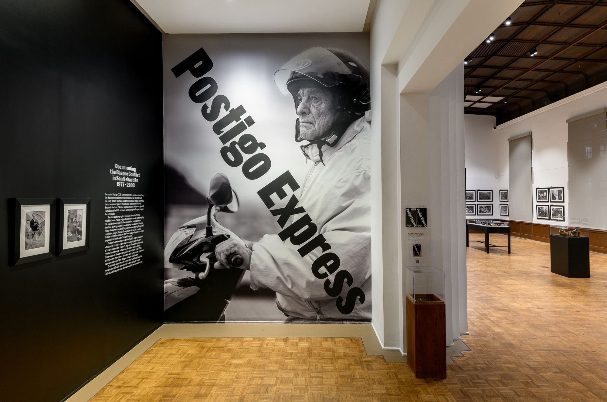 Entrance of white-walled gallery featuring huge wallpaper photo of elderly Postigo on a motorbike, behind huge diagonal title