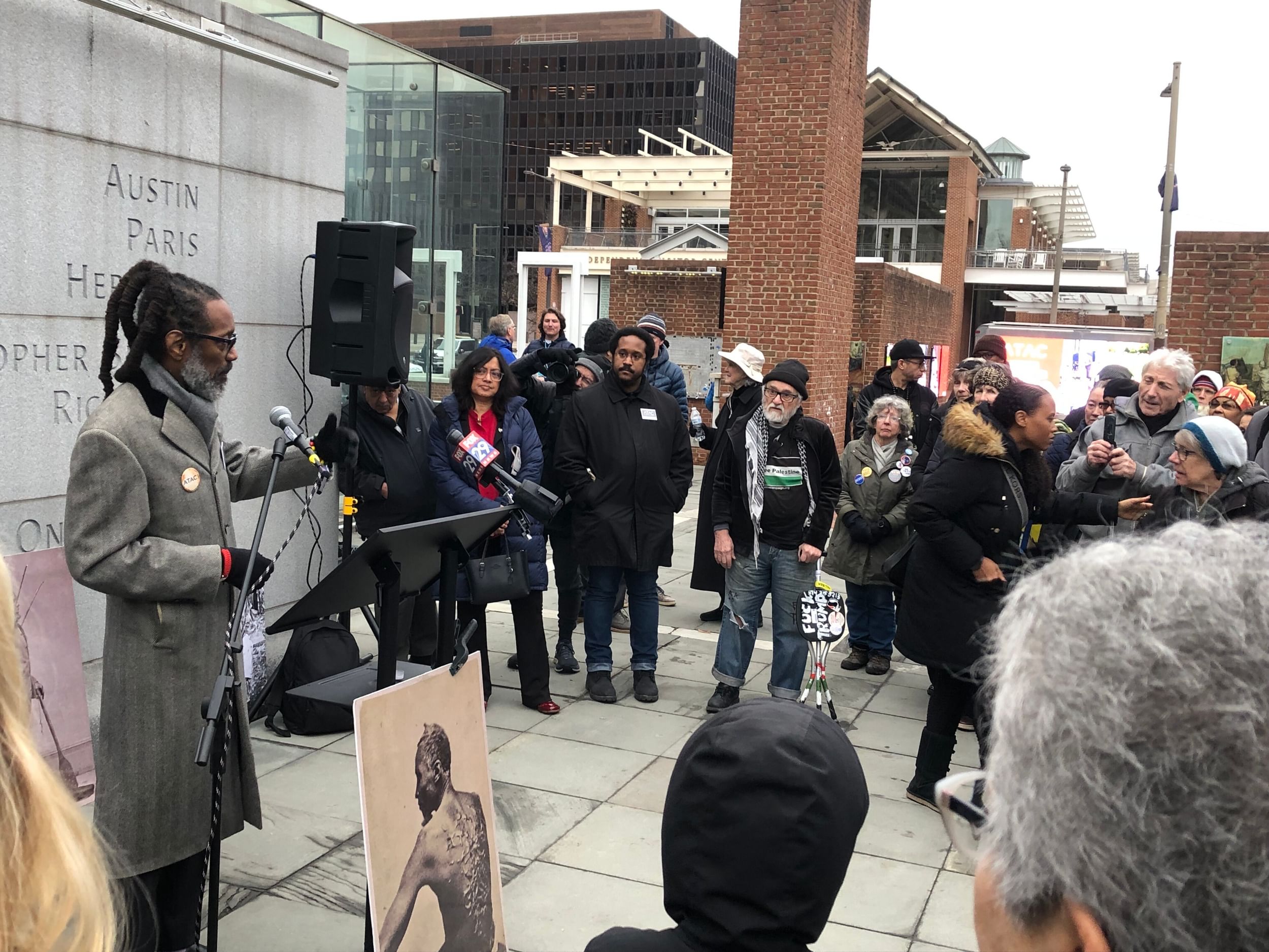 Coard, a Black man in a gray coat, speaks from a podium to a crowd of people outside on a winter afternoon.