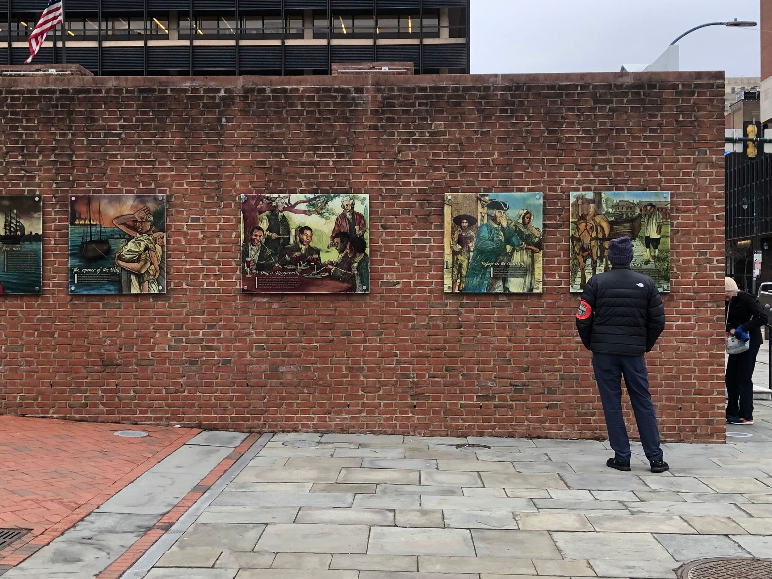 Seen from behind, a man looks at colorful square glass panels depicting a history of US slavery, mounted on a brick wall.