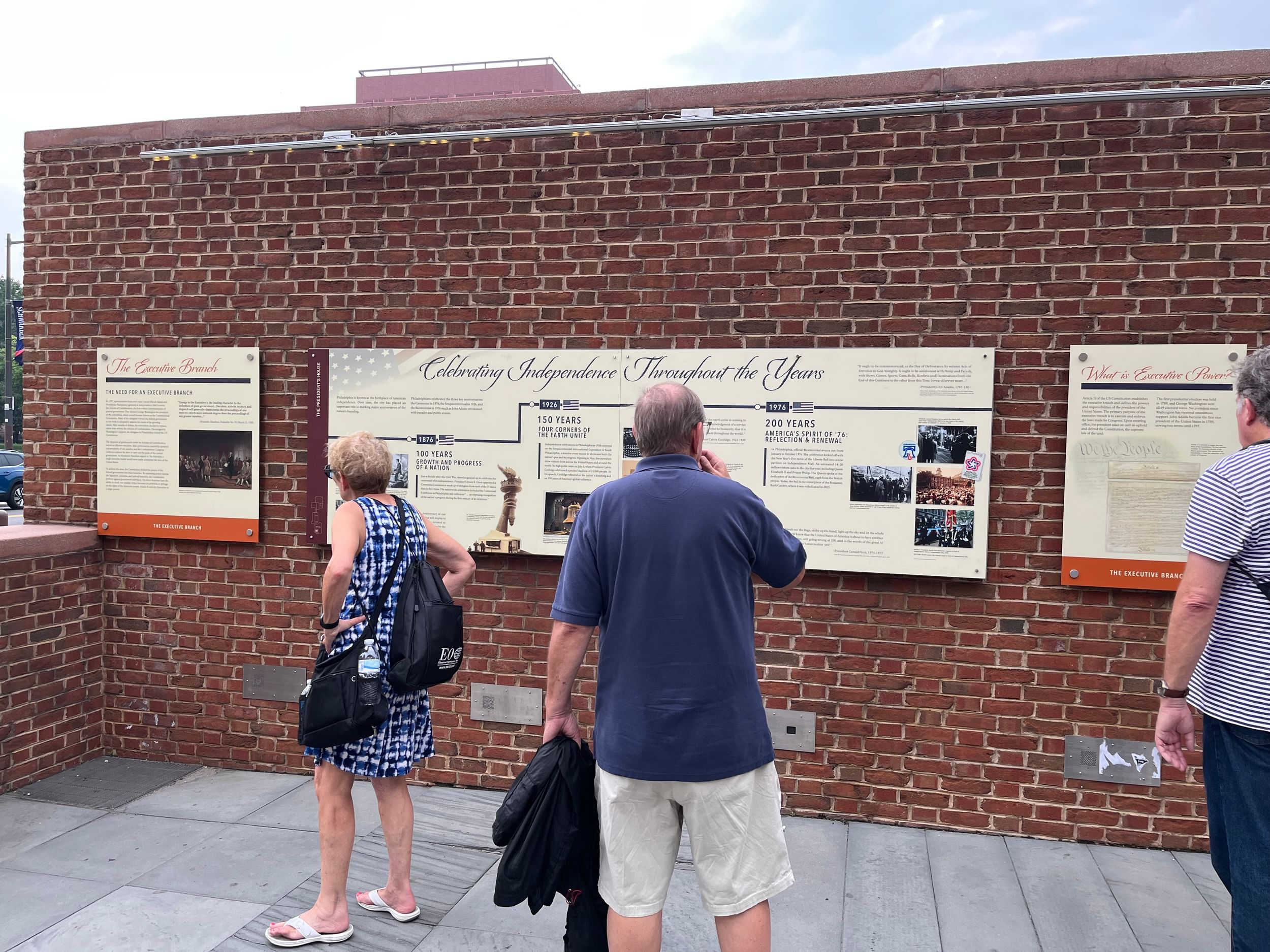 Three people in summer clothes look at beige panels with small text, mounted on a low brick wall.