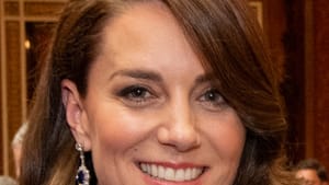 Close-up on smiling Kate. She wears diamond earrings and a high-necked blue dress, her brown hair curving around her face.