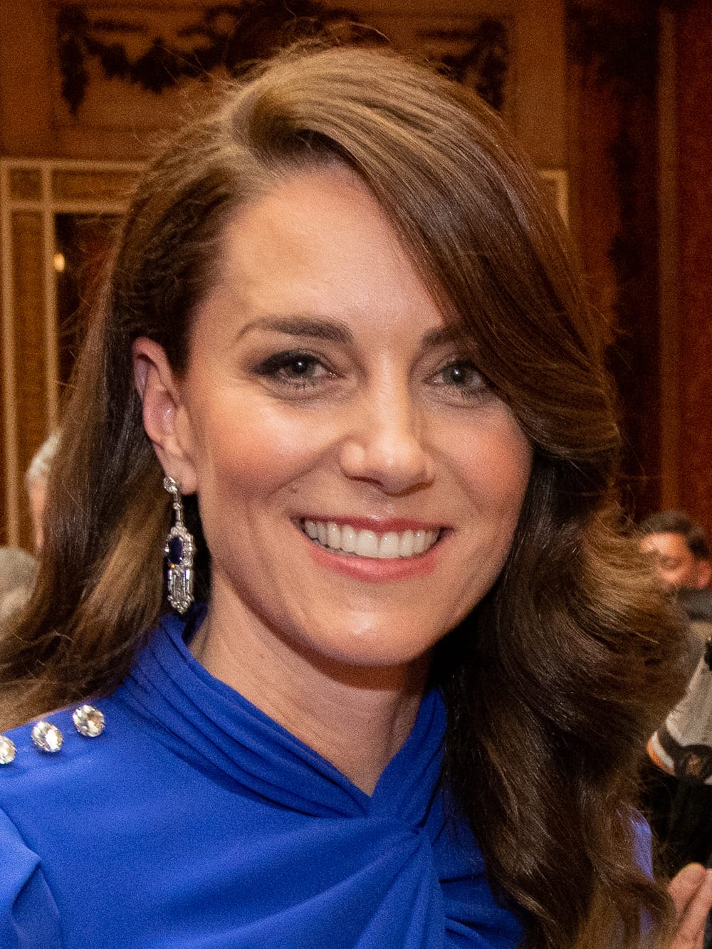 Close-up on smiling Kate. She wears diamond earrings and a high-necked blue dress, her brown hair curving around her face.