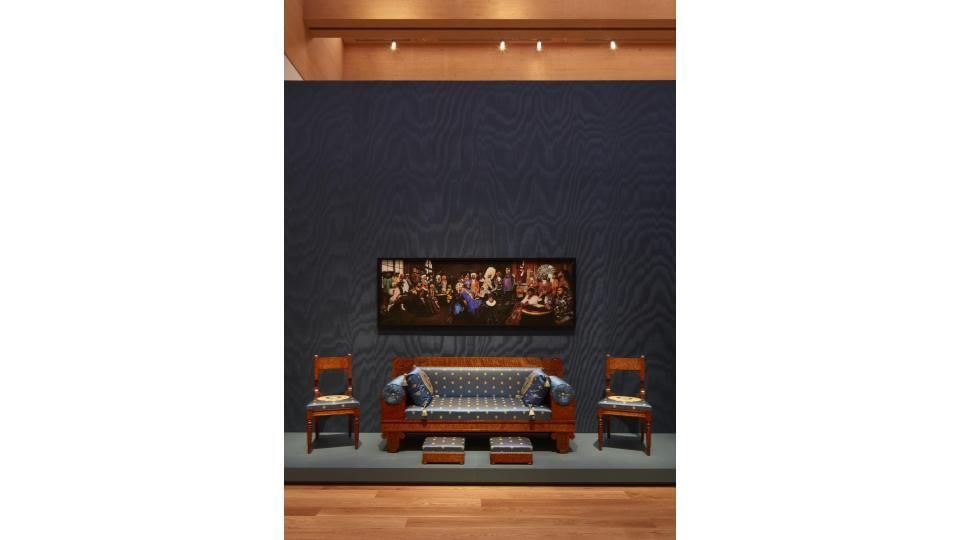 On a blue satin wall, Cox’s large photo, described in text below, above a blocky, ornate wooden sofa with blue silk cushions