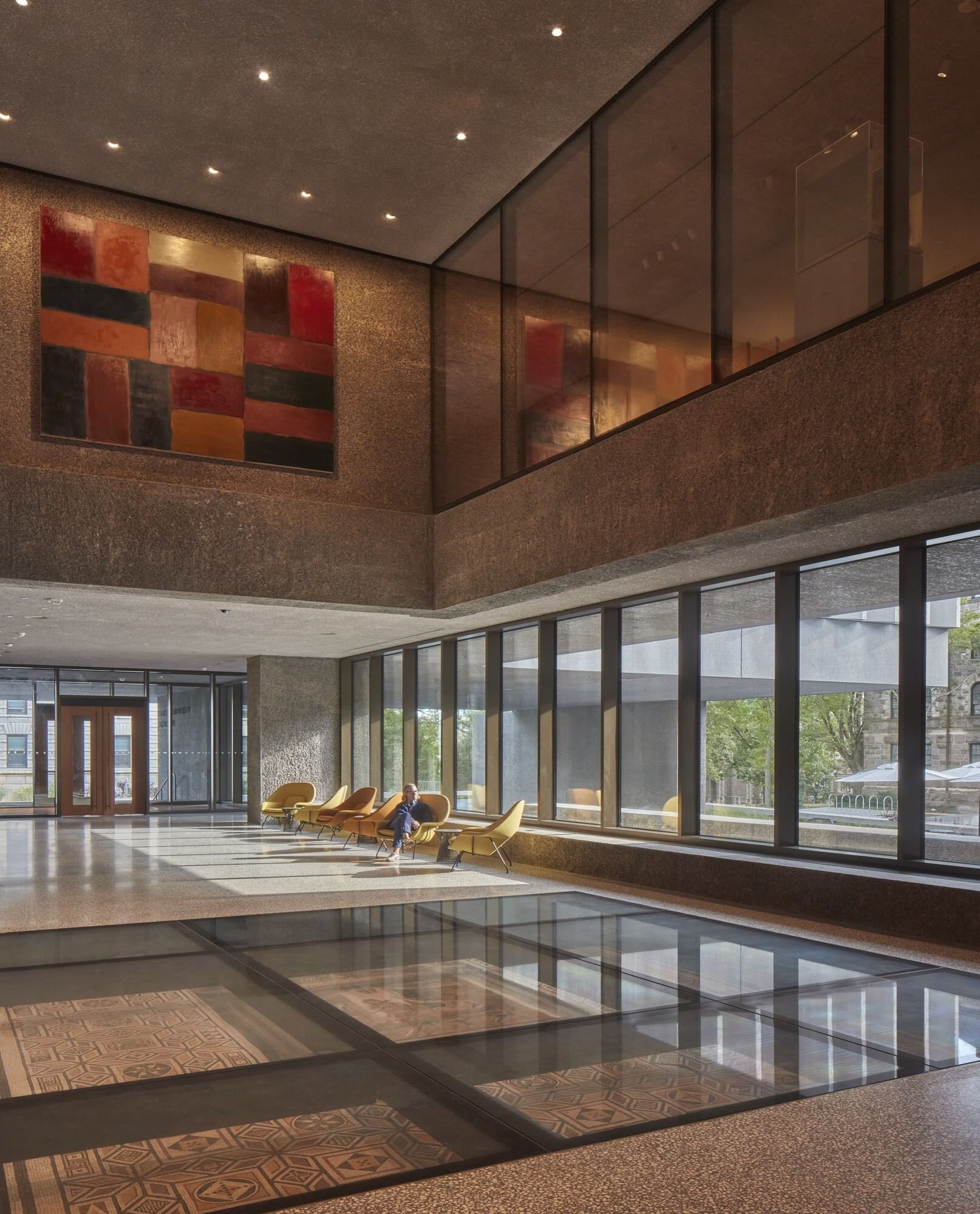 Interior view of a dynamic two-story stone hall with large rectangular windows and mosaics visible beneath glass in the floor