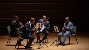 The four Black musicians, each wearing a blue or black suit, sit on chairs and play their instruments onstage.