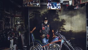 A Black man stands confidently in a garage with his custom motorcycle, against a backdrop of an 1800s Hudson Valley painting