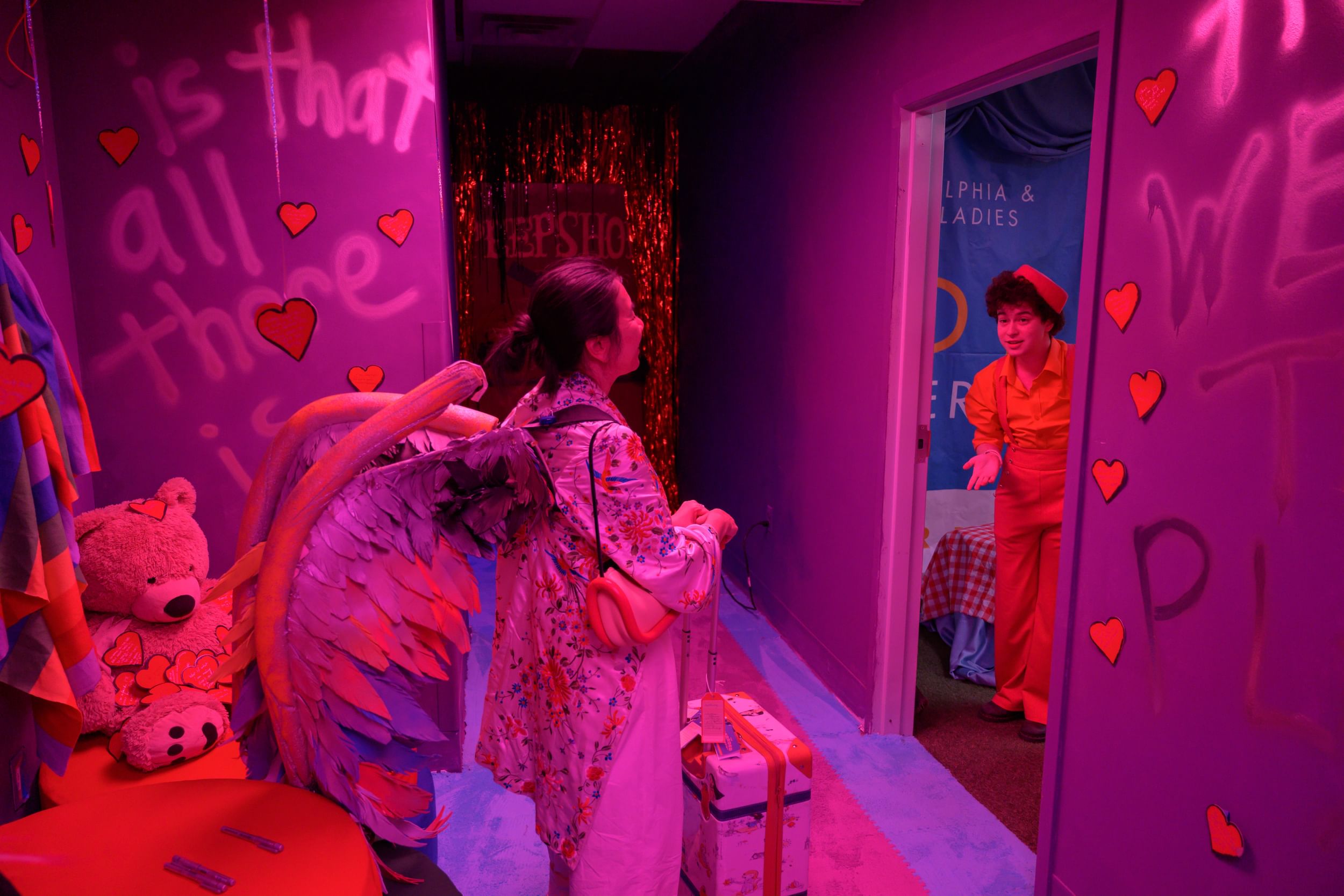 Two women stand in a pink-hued room is decorated with paper heart cutouts, streamers, and teddy bear.