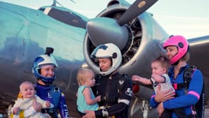 Three women in skydiving suits each hold one of three small children, an airplane parked behind them
